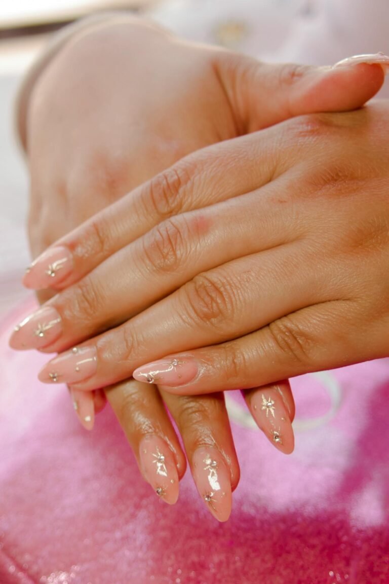 Close-up of a woman's hands showing elegant glitter nail art on a pink fabric background.